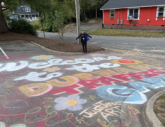 Photographed is a member of the Way to Go Durham team posing in front of a chalk filled parking lot. The chalk spells out “Welcome to the Durham Traffic Garden” in huge colorful letters surrounded by colorful flower drawings.